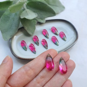 May include: A collection of teardrop-shaped glass beads with a pink and green ombre effect. The beads are displayed on a plate and in a hand.