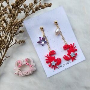 Puede incluir: Un par de pendientes de cangrejo rojo con una perla y un encanto de estrella de mar púrpura. Los pendientes están sobre un fondo blanco con un cortador de galletas de cangrejo rosa.
