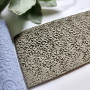 May include: A close-up of a brown clay rolling pin with a detailed embossed pattern. The pattern features a repeating design of circles, flowers, and swirls.