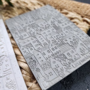 May include: A gray rubber stamp with a detailed design of a city skyline with houses, trees, and windows. The stamp is textured and has a raised design.