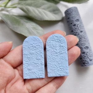 Può includere: Due orecchini in argilla polimerica blu chiaro con un motivo floreale. Gli orecchini hanno la forma di un rettangolo arrotondato con la parte superiore arrotondata.