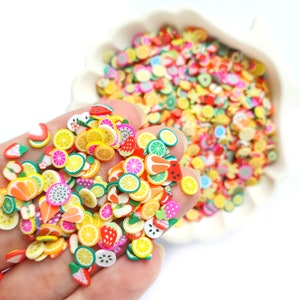 May include: A selection of colourful fruit-shaped polymer clay charms, including watermelon, lemon, lime, and strawberry slices. The charms are scattered on a white surface.