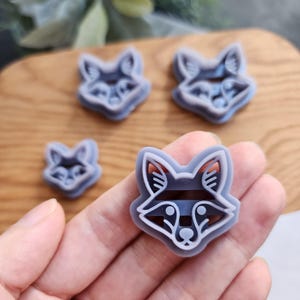 Puede incluir: Cuatro cortadores de galletas con forma de zorro de diferentes tamaños. Los cortadores son grises con contornos blancos, con diseños detallados de caras de zorro. Las caras de zorro tienen rayas y detalles de ojos. Los cortadores se sostienen en una mano.