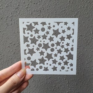 Puede incluir: Plantilla de plástico blanco con un patrón repetitivo de estrellas y círculos. La plantilla está diseñada para manualidades y decoración.