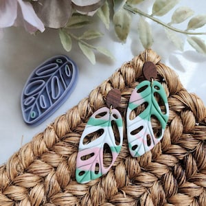 May include: Handmade polymer clay earrings in a Monstera leaf design, featuring a mix of green, pink, and white colors. The earrings have a wooden stud and are displayed with a leaf-shaped clay cutter on a woven surface.