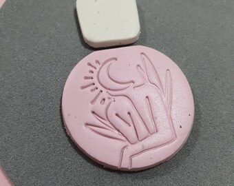 Puede incluir: Un sello de arcilla polimérica rosa con un diseño de una persona estilizada con una luna creciente sobre su cabeza y una planta detrás.