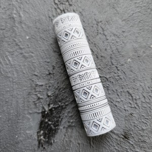 May include: A white cylinder with a geometric pattern. The pattern is made up of repeating squares, diamonds, and lines. The cylinder is about 5 inches long and 1 inch in diameter.
