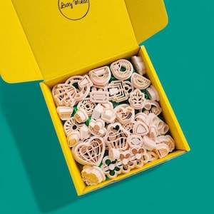 Puede incluir: Una caja amarilla con el texto "Lucy Molds" en la parte superior, abierta para revelar una variedad de cortadores de galletas de plástico rosa en diferentes formas, incluyendo corazones, estrellas, flores y formas geométricas.
