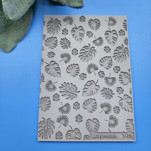 Puede incluir: Un molde rectangular gris con un patrón repetido de hojas tropicales, incluyendo diseños de monstera y palmera abanico. El molde está etiquetado "Luxymolds T-78" en la esquina inferior derecha. El fondo es azul liso.