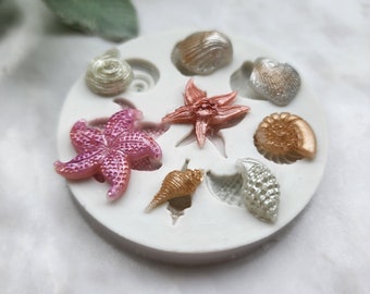 May include: A white silicone mold with eight cavities shaped like seashells and starfish. The mold is filled with pink, orange, and white resin casts of the shapes.