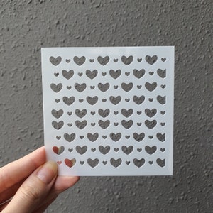 Puede incluir: Plantilla de plástico blanco con un patrón repetitivo de formas de corazón de diferentes tamaños. La plantilla está diseñada para manualidades y decoración.