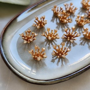 Puede incluir: Piezas de joyería doradas en forma de estrella, con pequeñas gemas transparentes. Las piezas están dispuestas sobre una bandeja de cerámica azul claro y gris. La joyería tiene una estética vintage o antigua.