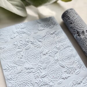 May include: A light blue rolling pin with a floral pattern embossed on the surface. The rolling pin is used for creating decorative patterns on fondant, clay, or dough.