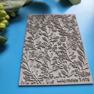 Puede incluir: Un sello de silicona gris con un diseño floral. El sello tiene un diseño en relieve de flores y hojas. El texto "Luxymolds T-176" está impreso en la esquina inferior derecha.