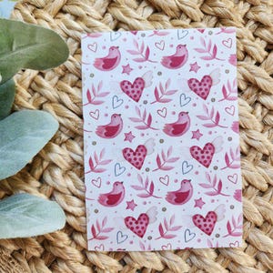 May include: A white background with a repeating pattern of pink birds, hearts, and leaves. The birds are stylised and have a simple design. The hearts are filled with a pink watercolour wash. The leaves are a light green.