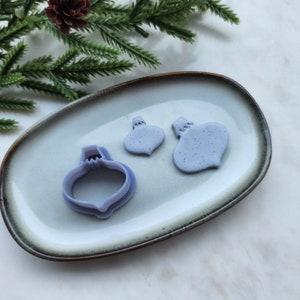 May include: Three purple plastic Christmas ornament cookie cutters on a light brown oval plate. The largest cutter is in the centre of the plate, and the two smaller cutters are to the right of the centre cutter.