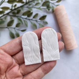 May include: Two white, arched-top clay earrings with a textured, wave-like pattern are held in a hand. A light pink, cylindrical object is in the background. Green leafy sprigs are also in the background.