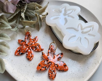 May include: Two pairs of orange and white speckled flower-shaped earrings with gold hoops. The earrings are next to a silicone mold of two flower shapes.