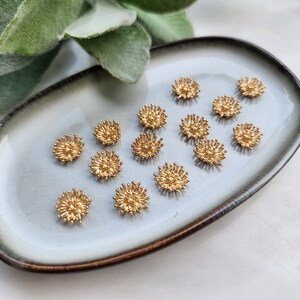 Puede incluir: Una colecci&oacute;n de 15 adornos met&aacute;licos de color dorado en forma de flor sobre un plato de cer&aacute;mica gris y marr&oacute;n. Los adornos tienen un dise&ntilde;o en relieve texturizado.