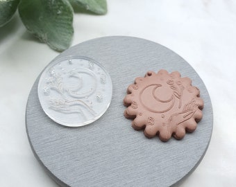 Puede incluir: Un sello de polímero transparente con un diseño de luna creciente, una mano y una rama de flores. El sello se utiliza para crear una impresión en arcilla, como se muestra en la imagen, un disco de arcilla marrón con el mismo diseño.
