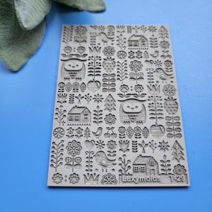 Puede incluir: Un sello de goma gris con un diseño de arte popular escandinavo con búhos, casas, flores y árboles. El sello está etiquetado como "Luxymolds T-29".