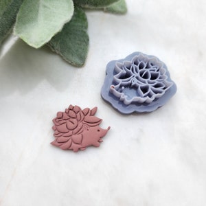 May include: A purple plastic cookie cutter in the shape of a flower and a brown plastic cookie cutter in the shape of a hedgehog.