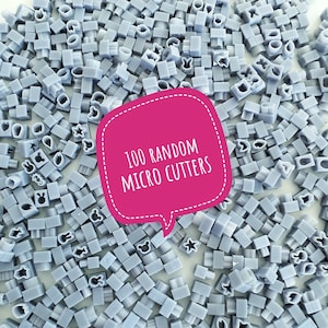 Puede incluir: Una imagen de primer plano de 100 microcortadores de plástico gris con varias formas. Los cortadores están dispuestos en un patrón aleatorio. La imagen incluye un globo de diálogo rosa con el texto "100 MICRO CORTADORES ALEATORIOS".