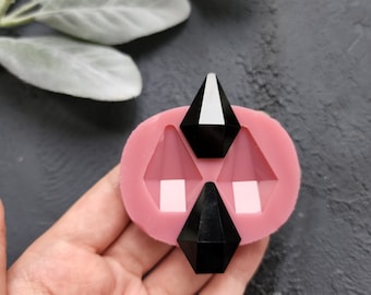 Puede incluir: Un molde de silicona rosa con tres formas geométricas y tres objetos negros facetados en forma de diamante. El molde es de color rosa claro y los objetos son negros con una superficie reflectante. El fondo es una superficie gris oscuro.