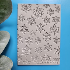 Puede incluir: Un molde de silicona gris con un diseño de múltiples formas de copo de nieve. El molde está etiquetado como "Luxymolds T-308".
