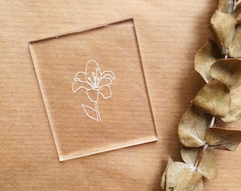 Puede incluir: Un sello de acrílico transparente con un contorno blanco de una flor de lirio.