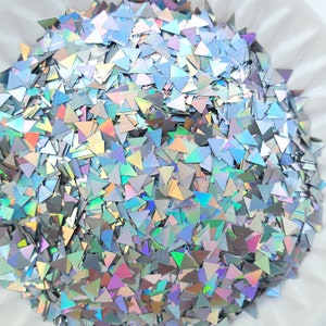 May include: A pile of iridescent triangle-shaped glitter. The glitter pieces are silver and reflect a rainbow of colors. The glitter is scattered and appears to be in a white bowl.