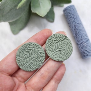 May include: A pair of green round earrings with a leaf pattern. The earrings are made of polymer clay and have a matte finish.