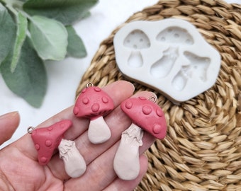 May include: Three red and white mushroom shaped polymer clay charms with gold jump rings. The charms are on a hand, and a silicone mold with three mushroom shapes is in the background.