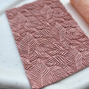 Puede incluir: Una estera de horneado de silicona rosa con un patrón de hojas en relieve. La estera está diseñada para usarse con fondant, pasta de goma y otras decoraciones comestibles.