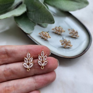 May include: A pair of gold-tone earring findings with a leaf design featuring clear rhinestones. The findings are shown on a hand and on a plate.