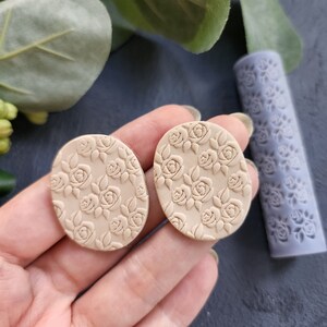 May include: Two beige oval-shaped polymer clay earrings with a floral design. The earrings are embossed with a repeating pattern of roses and leaves.