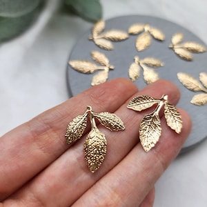 May include: Gold leaf charms with a textured finish. Each charm features three leaves connected by a stem. The charms have a hole at the top for attaching to jewelry or other crafts.