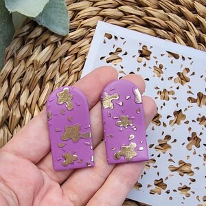 Puede incluir: Un par de pendientes en forma de arco de color lavanda con diseños de salpicaduras doradas. Los pendientes se muestran sobre un fondo de mimbre tejido y una plantilla blanca con un patrón floral.