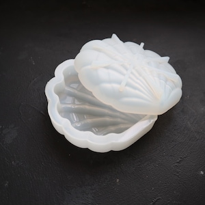 May include: A white silicone mold shaped like a seashell with a lid. The mold is open and the lid is sitting on top of the mold.