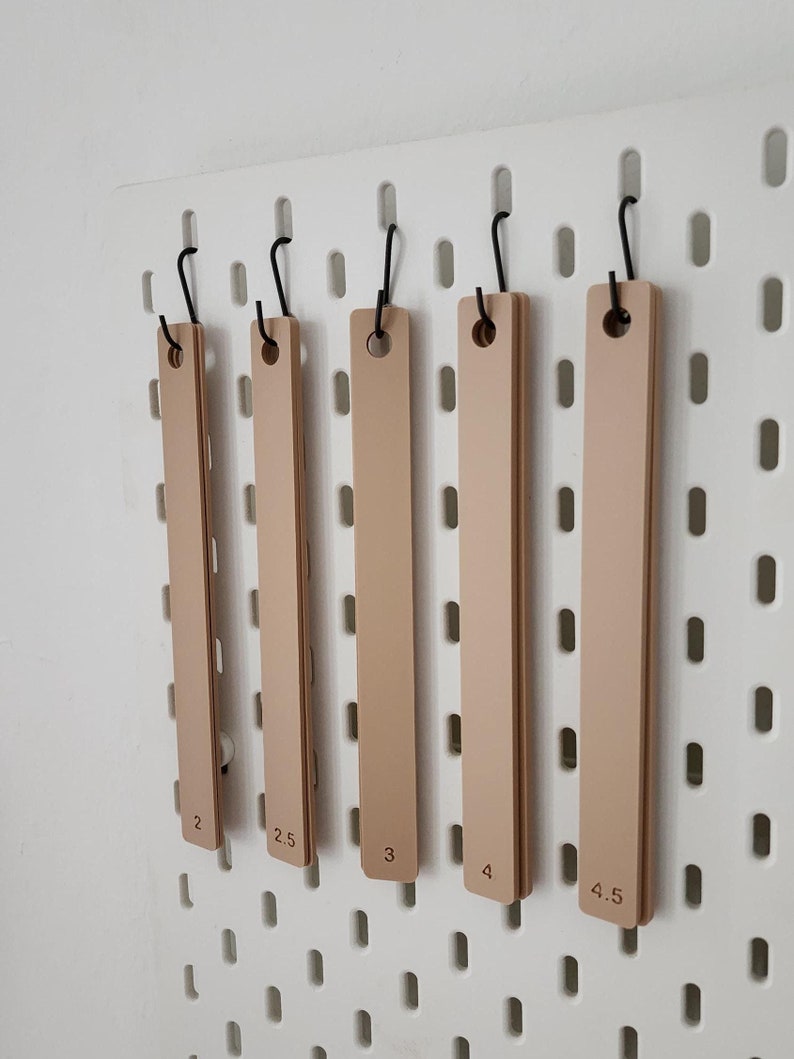 May include: Five beige wooden rulers hanging on hooks on a white pegboard. The rulers are labeled with measurements from 2 to 4.5.