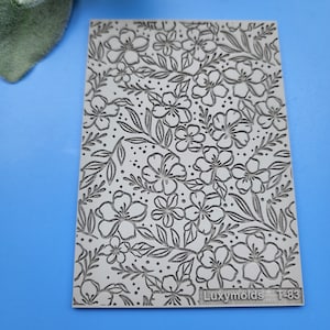 Puede incluir: Un primer plano de un molde de silicona gris con un patrón floral. El molde es texturizado y tiene un diseño en relieve de flores y hojas. El molde está etiquetado como "Luxymolds T-83".