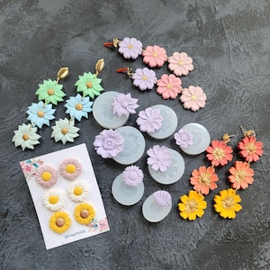 Puede incluir: Un surtido de pendientes y moldes con forma de flor de colores. Los pendientes son de varios colores, incluyendo amarillo, naranja, rosa y verde. Los moldes son de color azul claro y presentan diseños florales. La imagen también incluye una tarjeta con tachuelas en forma de flor.