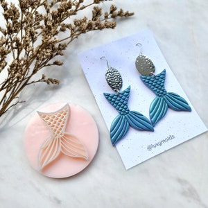 May include: A pair of mermaid tail earrings with silver-colored tops and blue tails. A pink circular mold with a mermaid tail design is also present. The earrings are displayed on a white card with the text "@luxymolds".