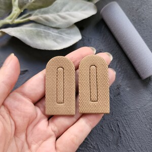 May include: A pair of brown arch-shaped earrings with a textured surface. The earrings are rectangular in shape with a cutout in the centre.