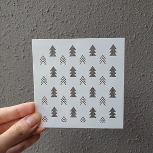 Puede incluir: Plantilla de plástico blanco con un patrón repetitivo de pequeños árboles de Navidad estilizados. La plantilla está diseñada para manualidades y decoración.