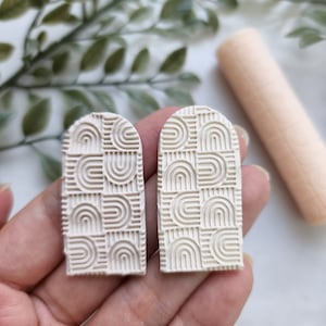 Può includere: Due orecchini in argilla polimerica di forma ad arco di colore avorio con un motivo geometrico di linee e archi in rilievo.