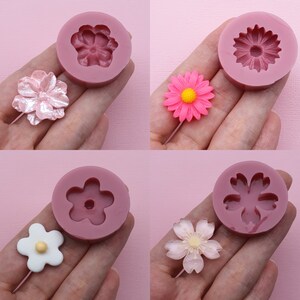 Könnte beinhalten: Vier Silikonformen zum Herstellen von blütenförmigen Dekorationen. Die Formen sind rosa und zeigen verschiedene Blumendesigns, darunter eine Gänseblümchen, eine Kirschblüte und eine einfache fünfblättrige Blume.