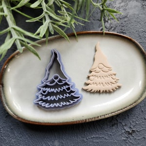 May include: Two plastic cookie cutters in the shape of a Christmas tree with a gnome face. The cutter on the left is purple and the cutter on the right is beige.