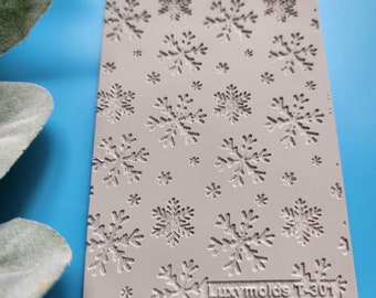 May include: A gray silicone mold with a snowflake pattern. The mold is textured and has the text "Lucy molds T-301" printed on the bottom right corner.