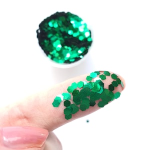 Può includere: Glitter verde a forma esagonale. Il glitter è sciolto e sparso su un dito.
